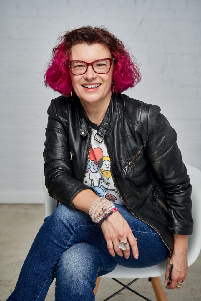 SpeakUp Founder Sharon Blady sitting in white chair with brick background. Sharon is wearing BT21 T-shirt, leather jacket and jeans.